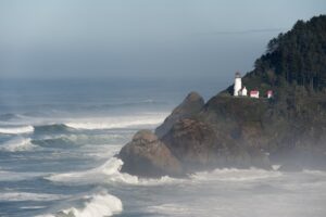 Crashing waves pound the rocky shore below a distant lighthouse in this horizontal landscape. The lighthouse sits atop a rugged, tree-covered headland as the ocean surges against the coastline. White waves roll into the beach, contrasting against the dark rocks and the bright white lighthouse.